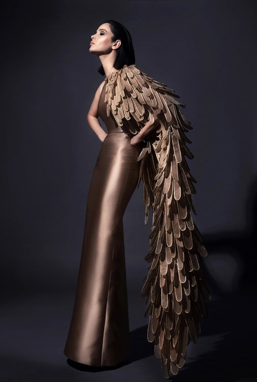 Sculpted one-shoulder gown in sandstone silk scuba and asymmetrical golden winged cape layered with 3D handcrafted plumes in crin and lamé